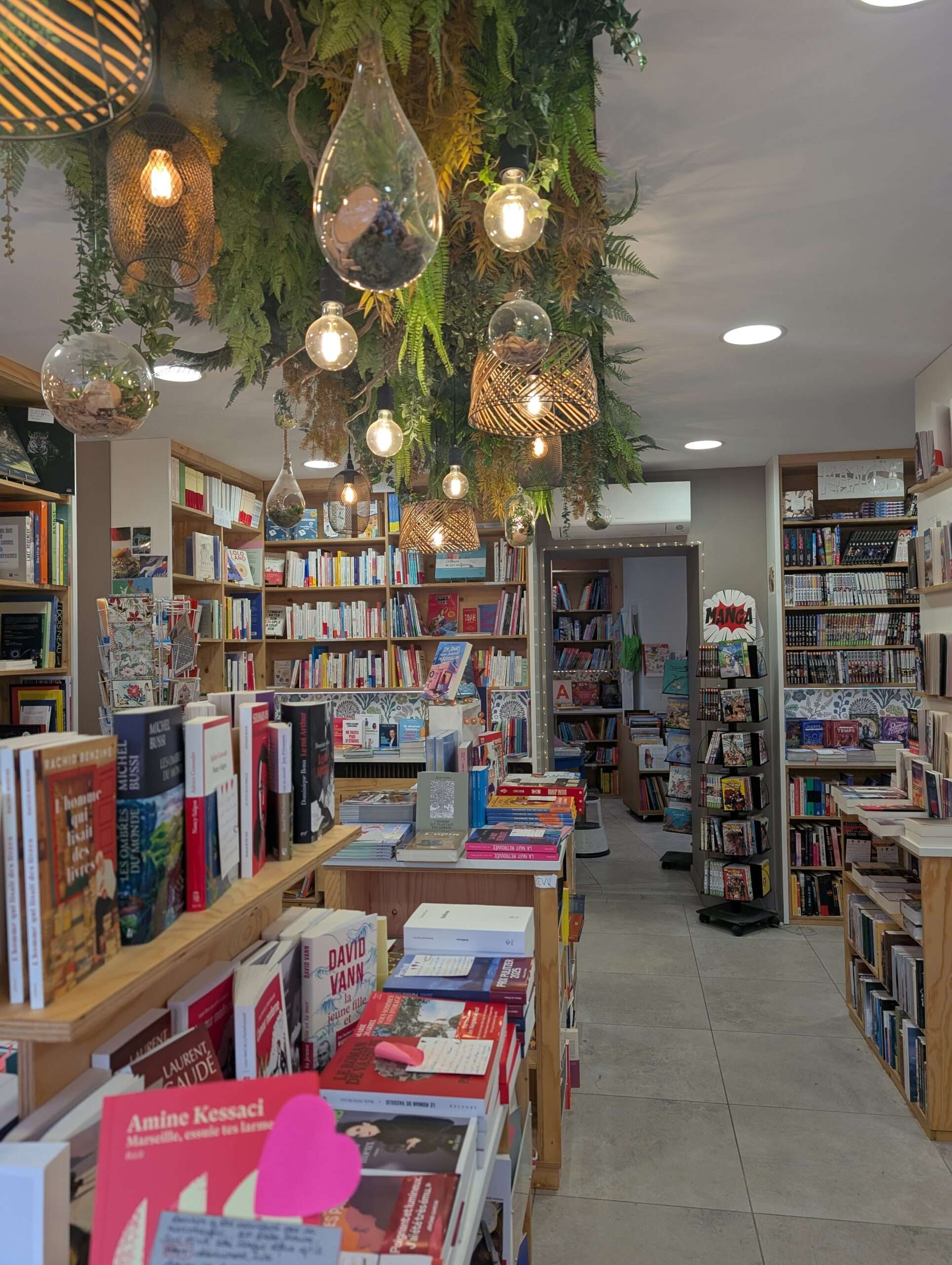 Intérieur de la librairie Le Jardin des Lettres à Craponne (69), lieu des ateliers d'écriture. Rayonnages en bois chargés de livres, tables de présentation avec titres en évidence. Décoration végétale suspendue et luminaires en verre — une atmosphère propice à l'écriture.